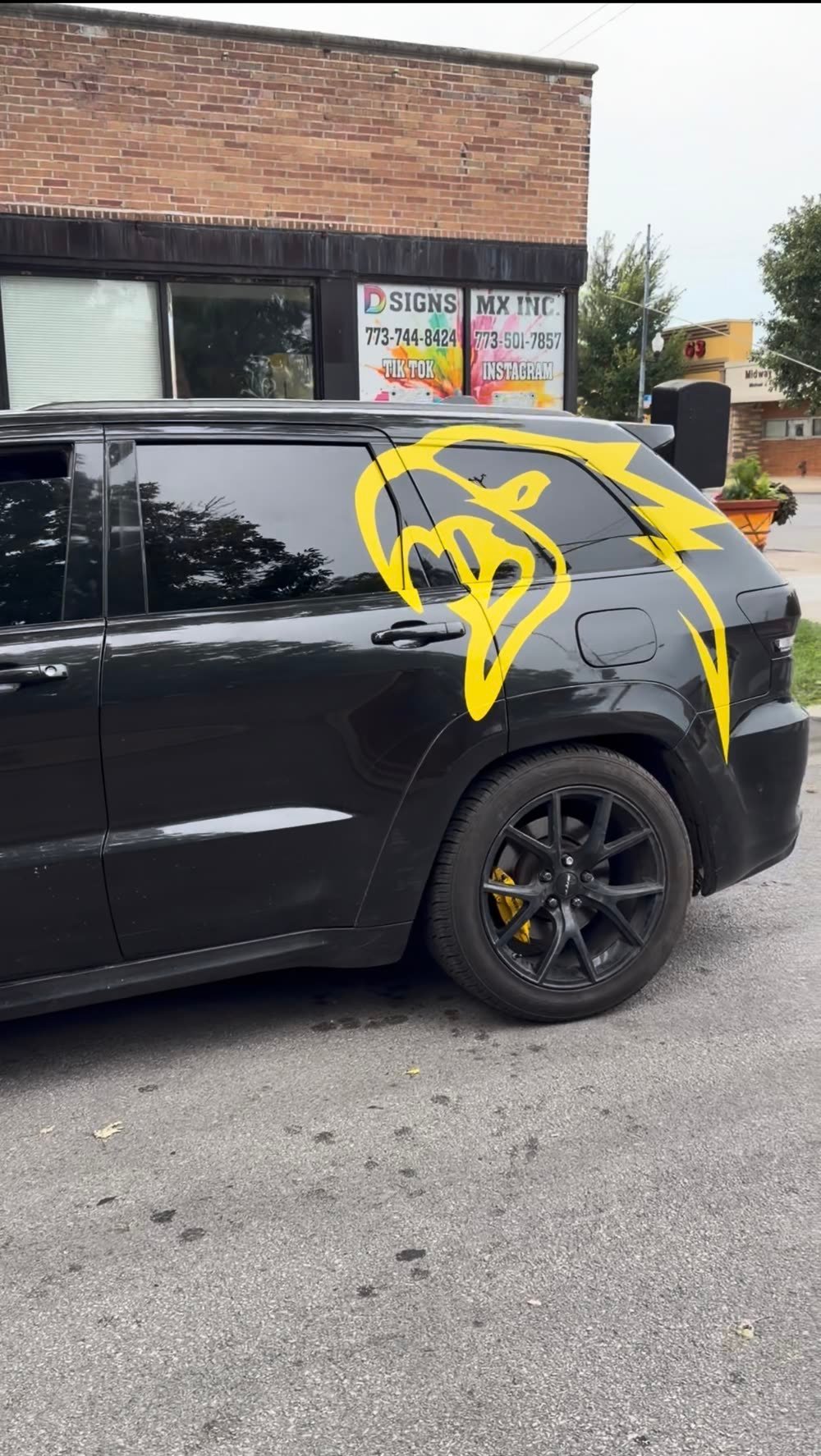Large yellow cat head decal on the rear side of a black SUV parked outside the DsignsMx shop